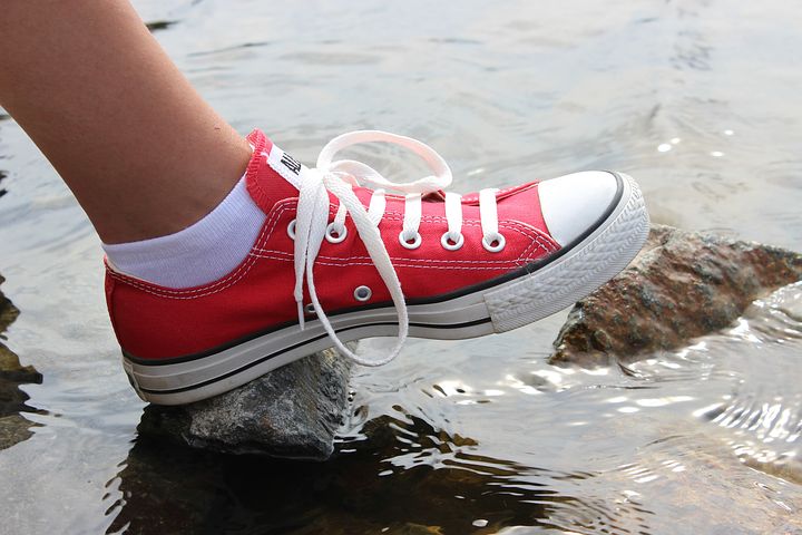 Das Bild zeigt einen Fuß in roten Turnschuhen, der auf einem im Wasser liegenden Stein steht.