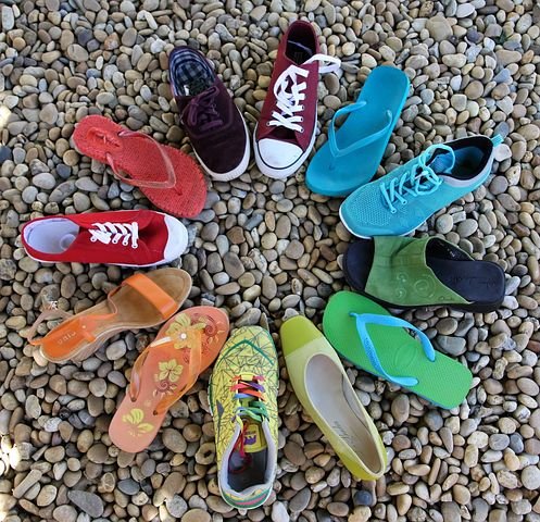 Zwölf unterschiedliche linke Schuhe (Turnschuhe, Sandalen, Pumps und Badelatschen) in den Farben grün, blau, rot, orange, gelb und lila stehen im Kreis angeordnet auf einem Kiesbett. 