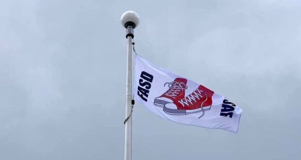 Auf dem Bild sieht man die Spitze eines Fahnenmastes. Daran flattert eine weiße Fahne im Wind. In der Mitte der Fahne sind zwei gemalte rote Turnschuhe zu sehen, an den Seiten steht FASD und SAFE.