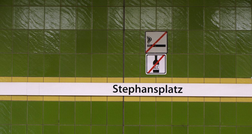 Das Bild zeigt eine grüne Fliesenwand. Darauf ist ein Schild mit der Aufschrift Stephansplatz angebracht. Über dem Schild kleben zwei Piktogramme an der Wand: Bitte nicht rauchen, bitte kein Alkohol.
