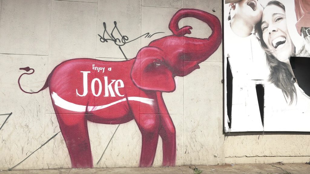 Auf die Außenwand eines Gebäudes ist ein roter Elefant gezeichnet. Auf seinem Bauch befindet sich ein weißer Schriftzug „Enjoy a Joke“, in Anlehnung an das Coca-Cola Flaschenlogo gestaltet. Rechts neben dem Elefanten sieht man einen Plakatausschnitt, auf dem zwei Menschen herzhaft lachen.