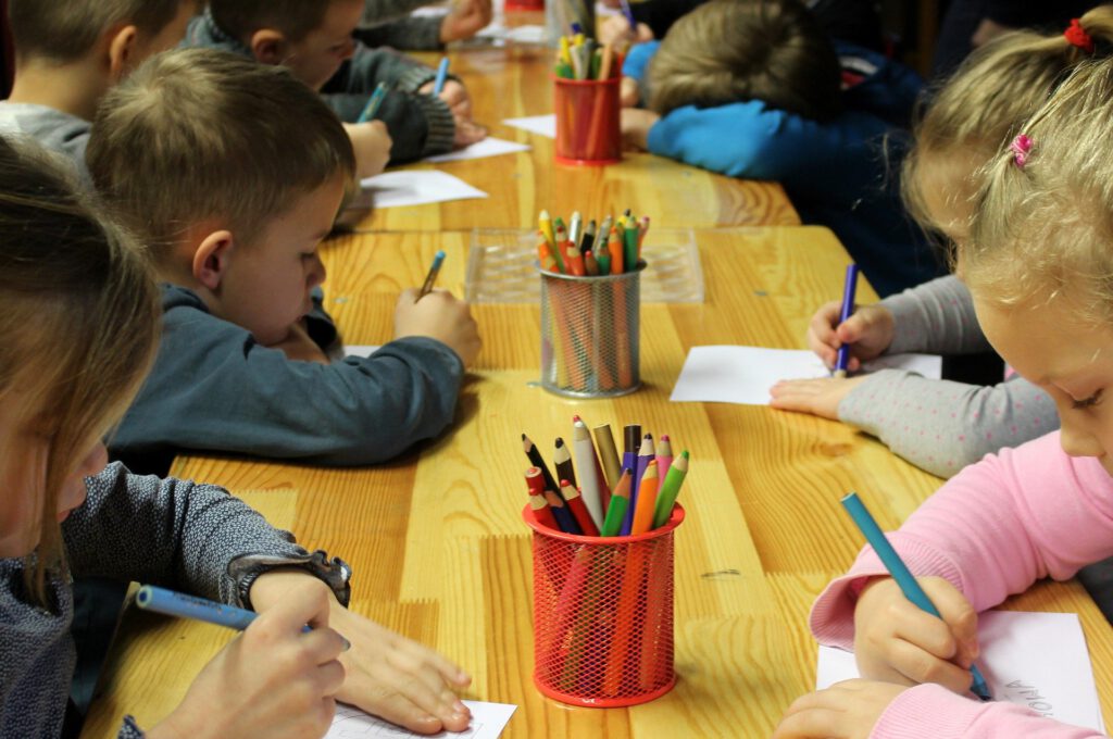 Jungen und Mädchen im Grundschulalter sitzen an einem langen Holztisch und malen. Auf dem Tisch stehen drei Becher mit Buntstiften.