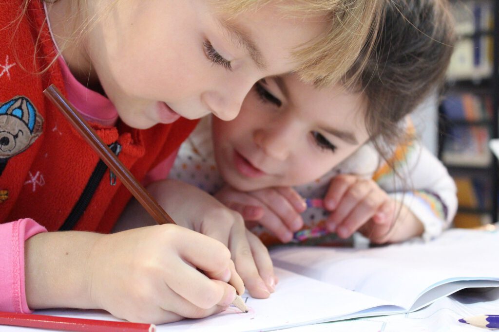 Auf dem Bild sieht man zwei Mädchen im Grundschulalter, die sich gemeinsam über ein Heft beugen. Eins der Mädchen schreibt bzw. malt mit einem Bleistift in das Heft.