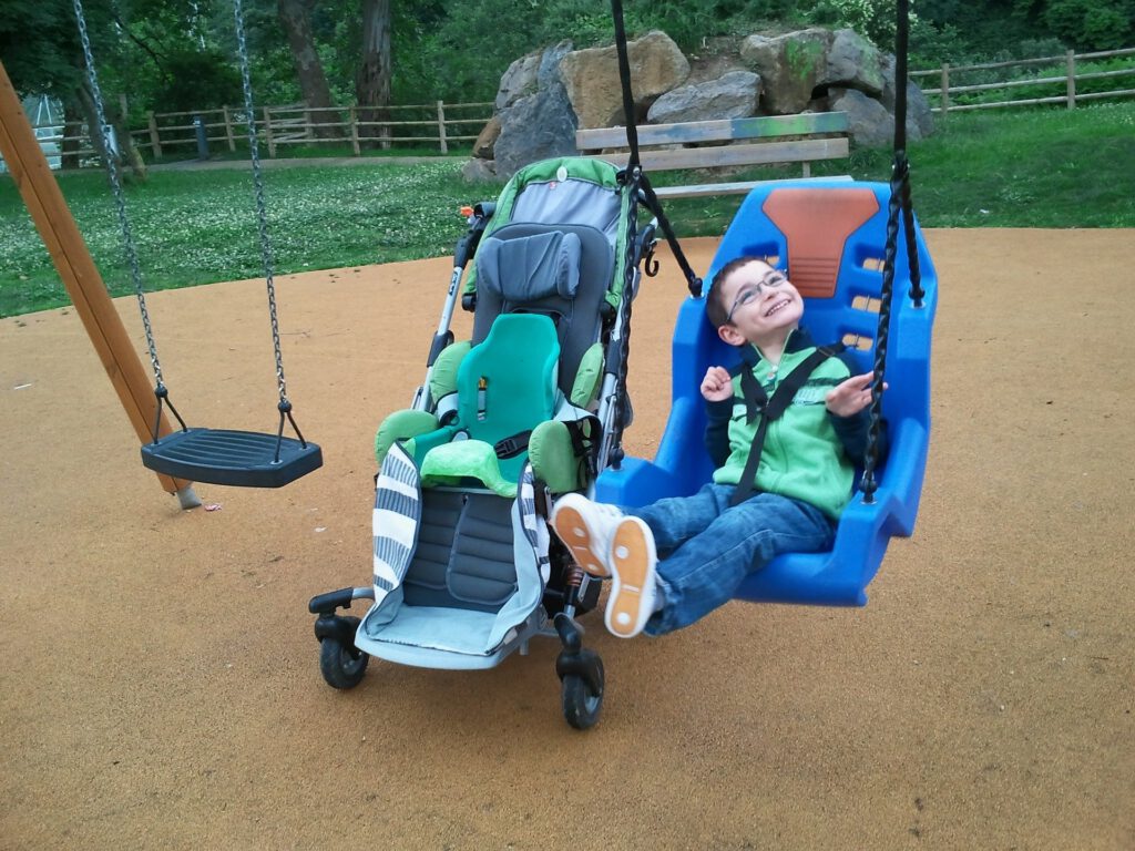 Das Bild zeigt einen körperbehinderten Jungen auf einer behindertengerechten Schaukel. Links von ihm steht sein Buggy. Daneben hängt eine normale Schaukel.