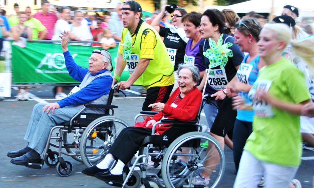 Das Bild zeigt mehrere Läuferinnen und Läufer eines Volkslaufs, die einen alten Mann und eine alte Frau im Rollstuhl beim Laufen vor sich her schieben.