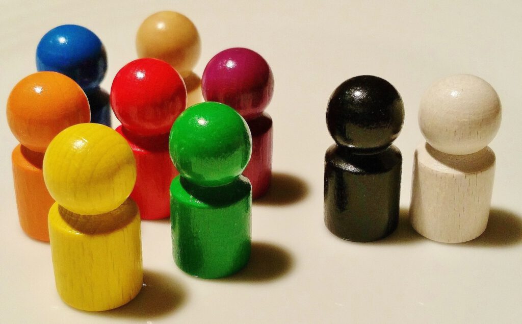 Auf der linken Seite des Bildes sieht man eine Gruppe bunter Spielfiguren aus Holz, die im Kreis zusammen stehen. Auf der rechten Bildseite stehen eine schwarze und eine weiße Spielfigur nebeneinander, mit deutlichem Abstand zu den andern.