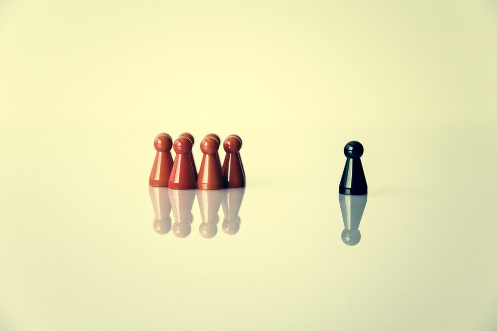 Auf der linken Seite des Bildes sieht man eine Gruppe roter Mensch-Ärger-Dich-Nicht-Figuren eng nebeneinander stehend. Auf der rechten Bildseite steht eine einzelne schwarze Spielfigur, mit deutlichem Abstand zu den andern.