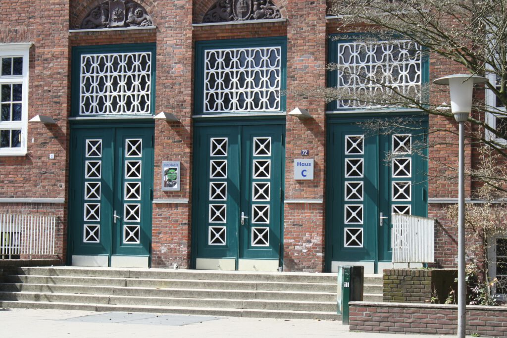 Das Bild zeigt den Eingang einer Schule in Hamburg: Treppenstufen führen zu drei großen grünen Türen mit weißen Verzierungen.