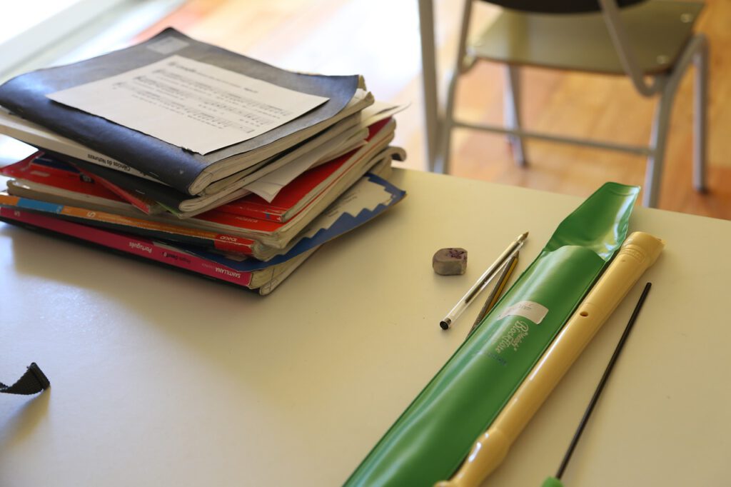 Auf einem Schultisch liegt ein Stapel Hefte. Zuoberst erkennt man ein Notenblatt. Rechts neben dem Stapel liegen ein Radiergummi, ein Kuli, ein Bleistift, eine grüne Flötenhülle und eine helle Blockflöte.