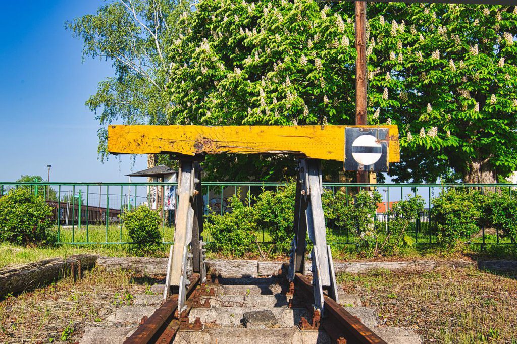 Das Bild zeigt das Ende eines Stumpfgleises, gesichert mit einem Prellbock. Im Hintergrund sieht man eine blühende Kastanie und Bahnanlagen.