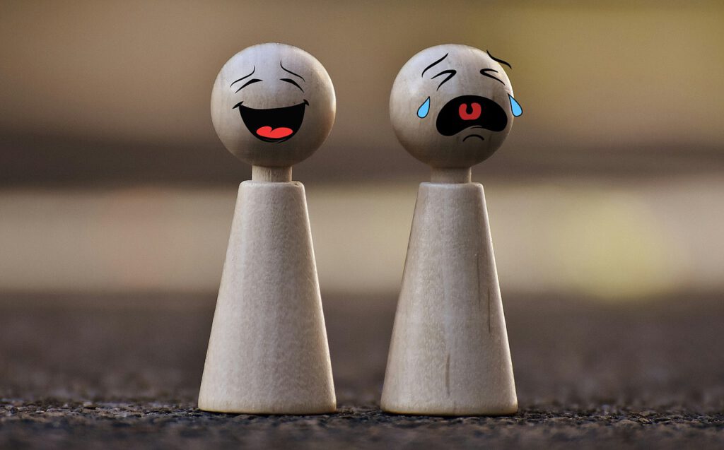 Auf dem Bild sieht man zwei kleine Spielfiguren mit aufgemalten Gesichtern. Die linke Spielfigur hat ein lachendes Gesicht, die rechte ein weinendes.