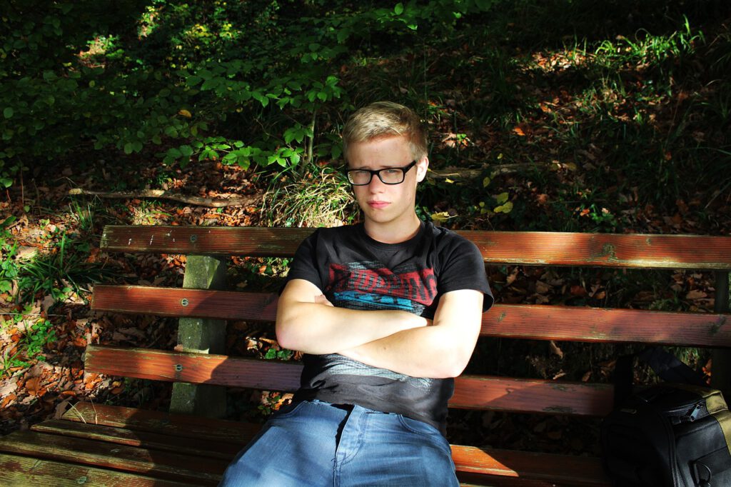 Ein junger Mann sitzt auf einer Holzbank und blickt skeptisch Richtung Kamera. Im Hintergrund sieht man Blätter und Büsche.