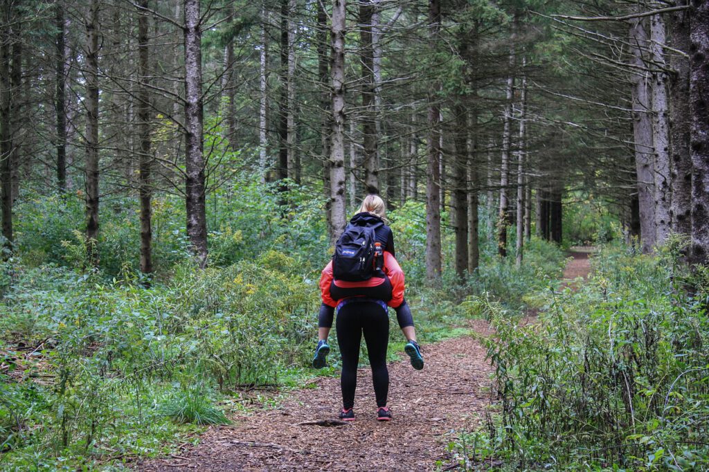 Das Bild zeigt eine Frau, die ein älteres Kind huckepack durch einen Wald trägt. Man sieht beide von hinten. Das Kind trägt einen großen  Rucksack.