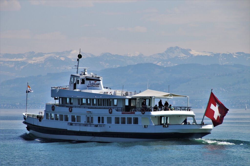 Das Bild zeigt ein weißes Ausflugsboot auf einem See.
Am Bug des Bootes weht die Flagge der Schweiz. Im Hintergrund sieht man schneebedeckte Berggipfel.