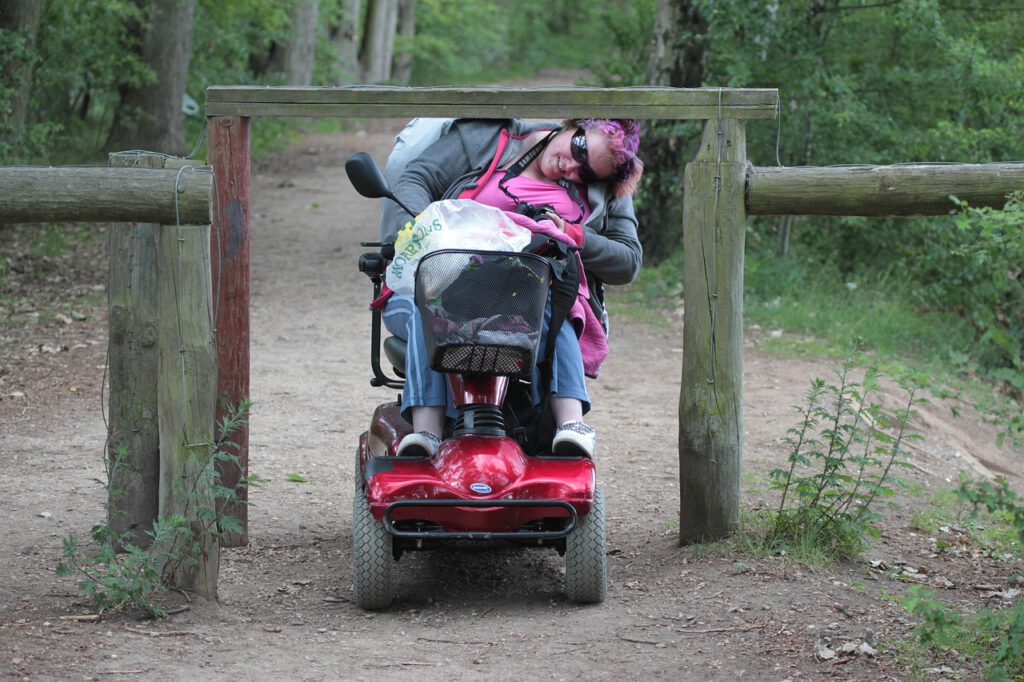 Das Bild zeigt eine junge Frau im Elektro-Rollstuhl. Die Frau muss sich bücken, um mit ihrem Rollstuhl durch ein Geländer aus Holz hindurchfahren zu können.