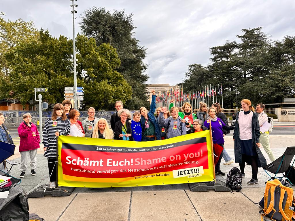 Eine Gruppe von Menschen steht hinter einem großen schwarz-rot-gelben Banner mit der Aufschrift: "Seit 14 Jahren gilt die UN-Behindertenrechtskonvention: Schämt Euch! Shame on you! Deutschland verweigert das Menschenrecht auf inklusive Bildung. Eltern fordern Inklusion. Jetzt endlich!" Im Hintergrund sieht man den Uno-Hauptsitz in Genf.