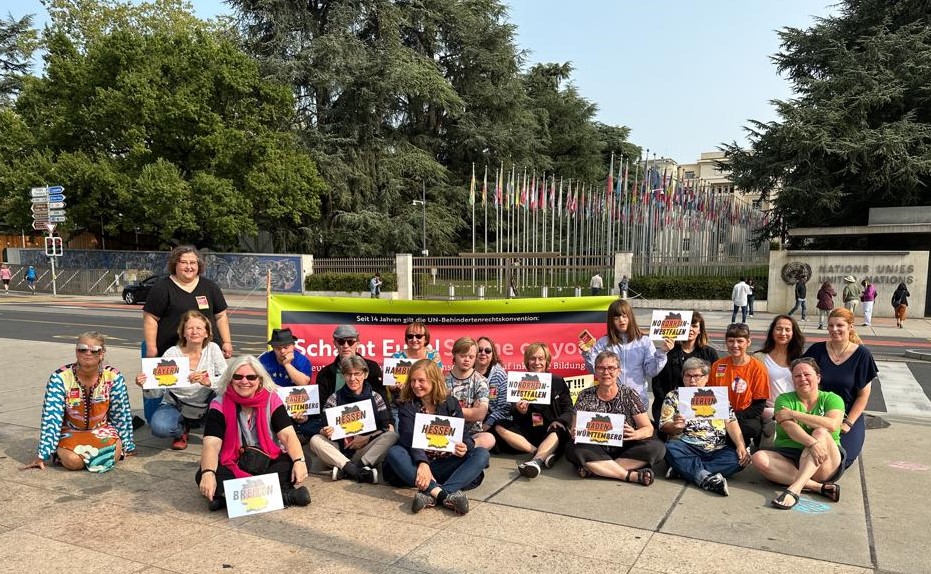 Menschen sitzen im Schneidersitz auf dem Boden eines gepflasterten Platzes. Mehrere halten Schilder mit den Namen verschiedener Bundesländer in den Händen. Im Hintergrund sieht man ein großes Banner und die Fahnenallee vor dem Uno-Hauptsitz in Genf.