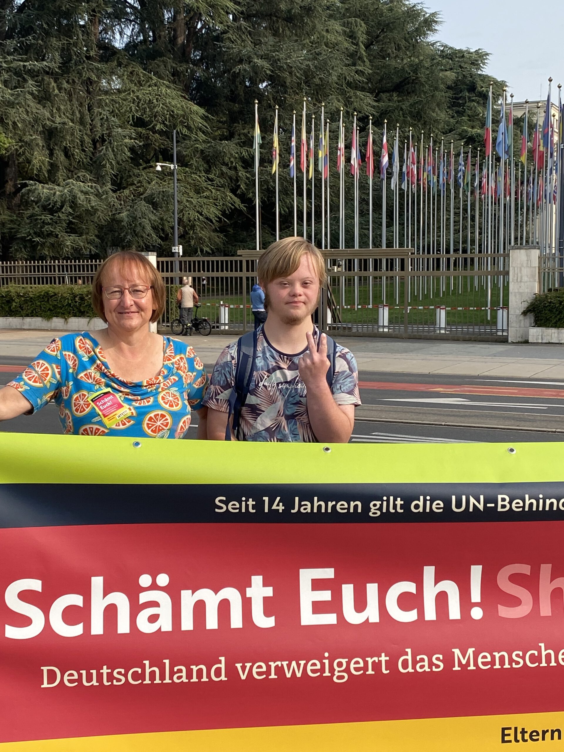 Auf dem Bild sieht man einen jungen Mann und mich hinter unserem Protest-Banner "Schämt Euch". Im Hintergrund erkennt man Teile der Fahnenallee vor dem Uno-Hauptsitz in Genf.