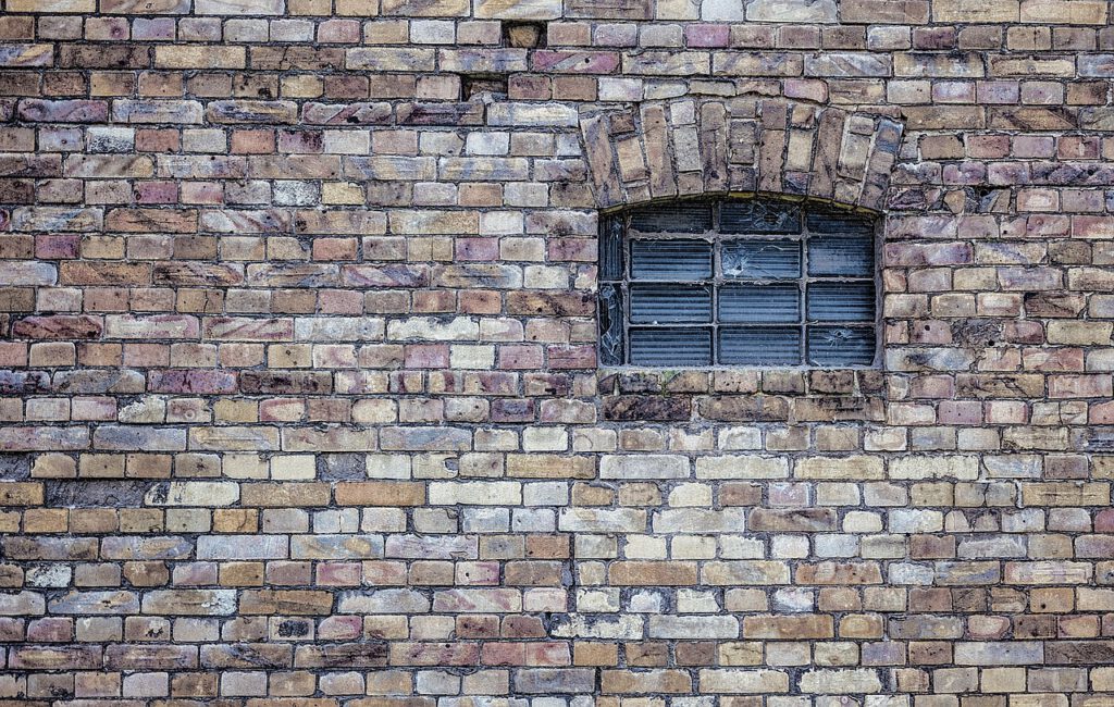 Das Bild zeigt eine alte Hauswand, gemauert aus rot-braunen Ziegeln. Rechts in der Wand sieht man ein kleines, vergittertes Fenster.
