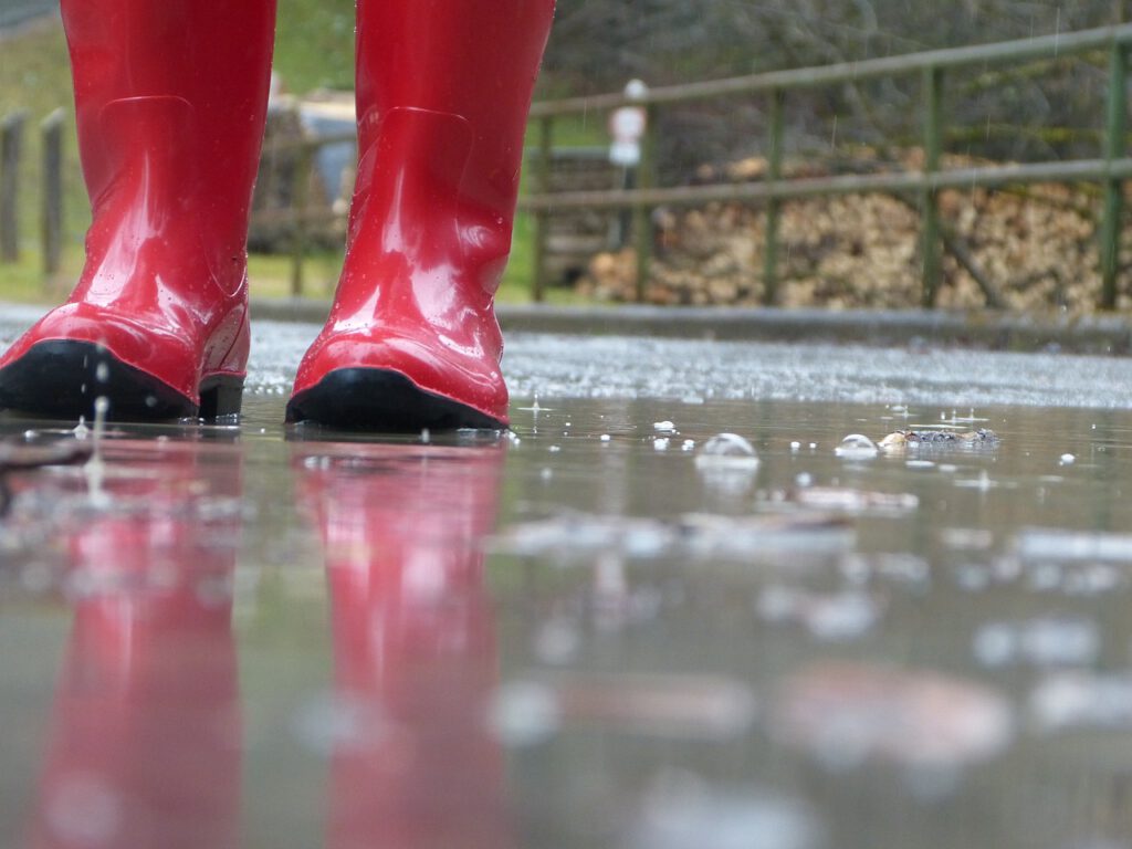 Das Bild zeigt ein Paar rote Gummistiefel, die im Regen in einer Pfütze auf einer Brücke stehen.