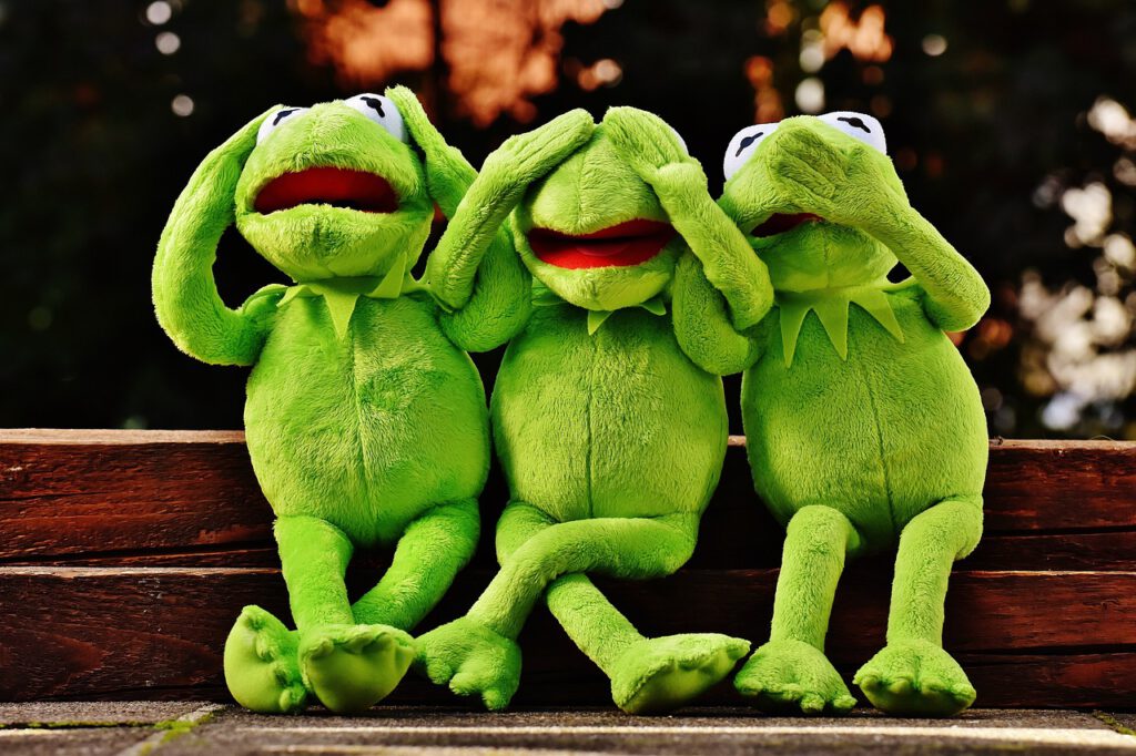 Drei grüne Kermit-Puppen sitzen nebeneinander auf zwei Holzbalken. Die erste Frosch-Figur hält sich die Ohren zu. Die zweite Frosch-Figur hält sich die Augen zu. Die dritte hält sich den Mund zu.