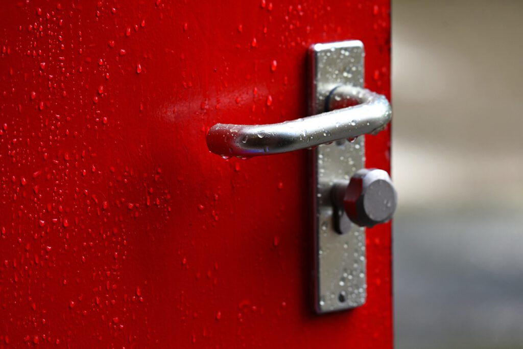 Auf dem Bild sieht man einen Teil einer geöffneten roten Tür mit silbernem Türgriff. Auf Griff und Tür sind Wassertropfen.