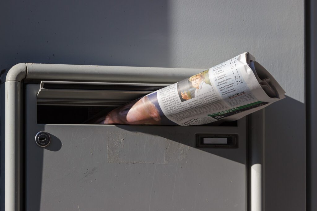 Auf dem Bild sieht man den oberen Teil eines grauen Briefkastens. In der Klappe des Briefkastens steckt eine zusammengerollte Zeitung.