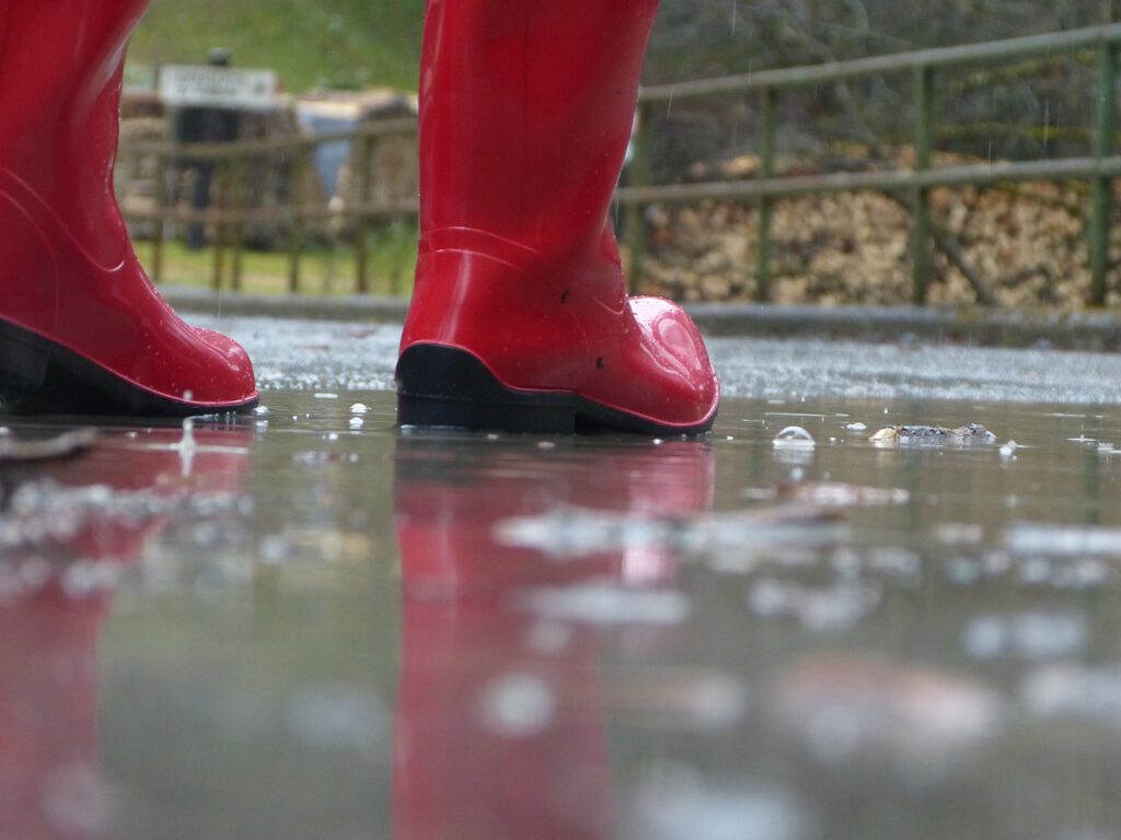 Zwei rote Gummistiefel spiegeln sich in einer großen Pfütze mit Regentropfen.