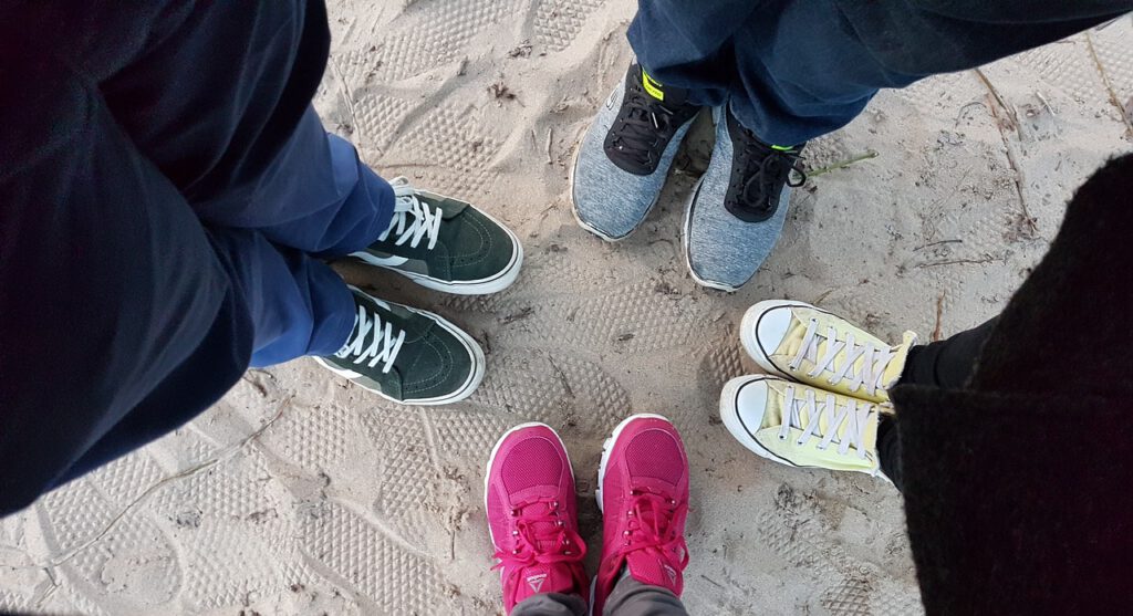 Vier Menschen stehen in einem Kreis zusammen. Das Bild zeigt den Blick auf  ihre Beine und Füße. Alle tragen dunkle lange Hosen und Turnschuhe in unterschiedlichen Farben. 