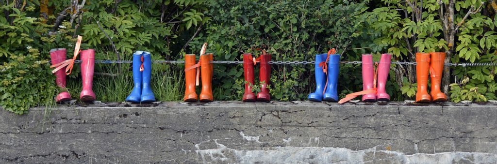 Auf dem Bild sieht man 7 Paar Gummistiefel, die auf einer Steinmauer aufgereiht stehen. Zwei Paar Gummistiefel sind pink, zwei Paar blau, zwei Paar orange und ein Paar rot. Hinter der Mauer und den Gummistiefeln sieht man grüne Büsche und Bäume.