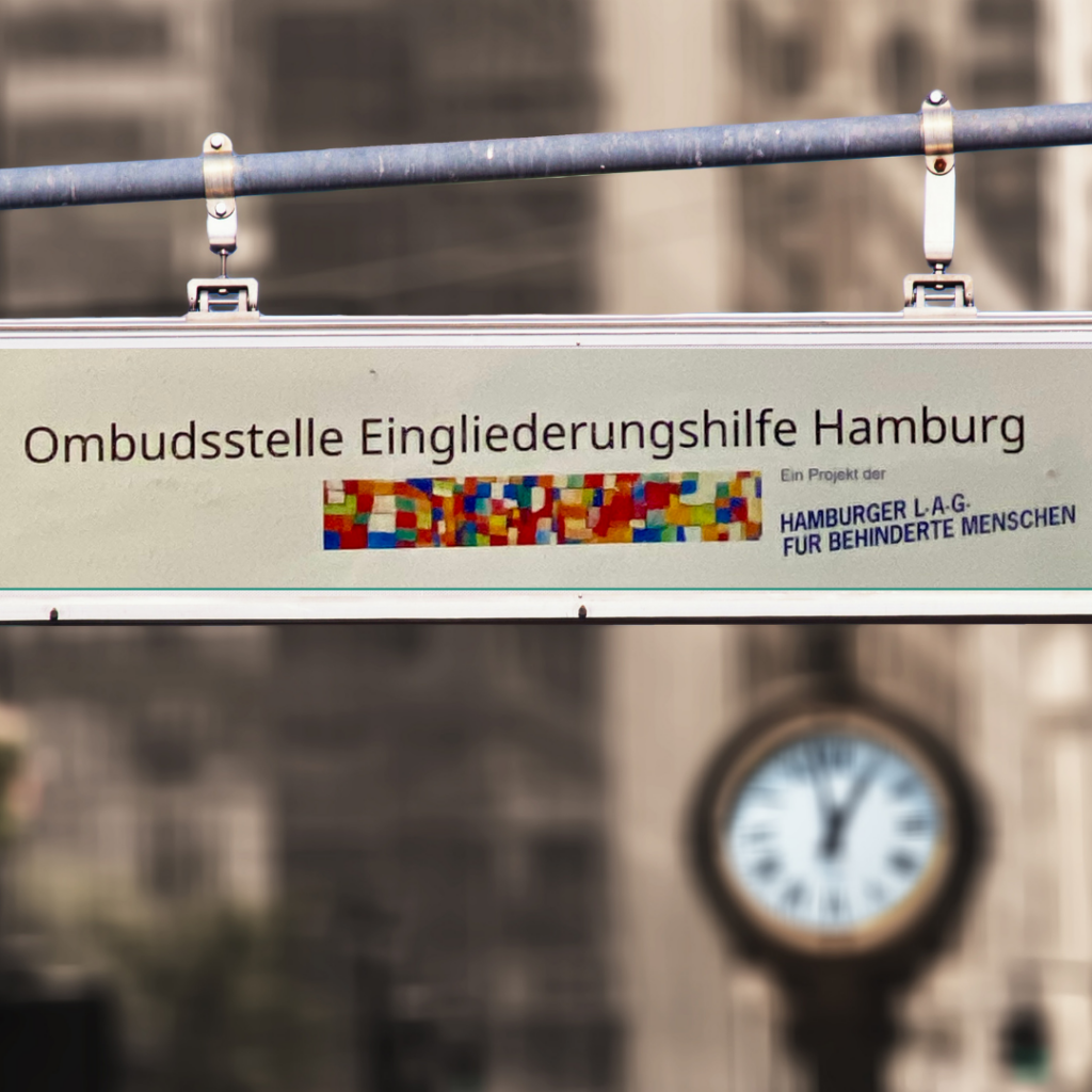 Das Bild zeigt ein Schild mit der Aufschrift "Ombudsstelle Eingliederungshilfe Hamburg. Ein Projekt der Hamburger L.A.G. für behinderte Menschen"