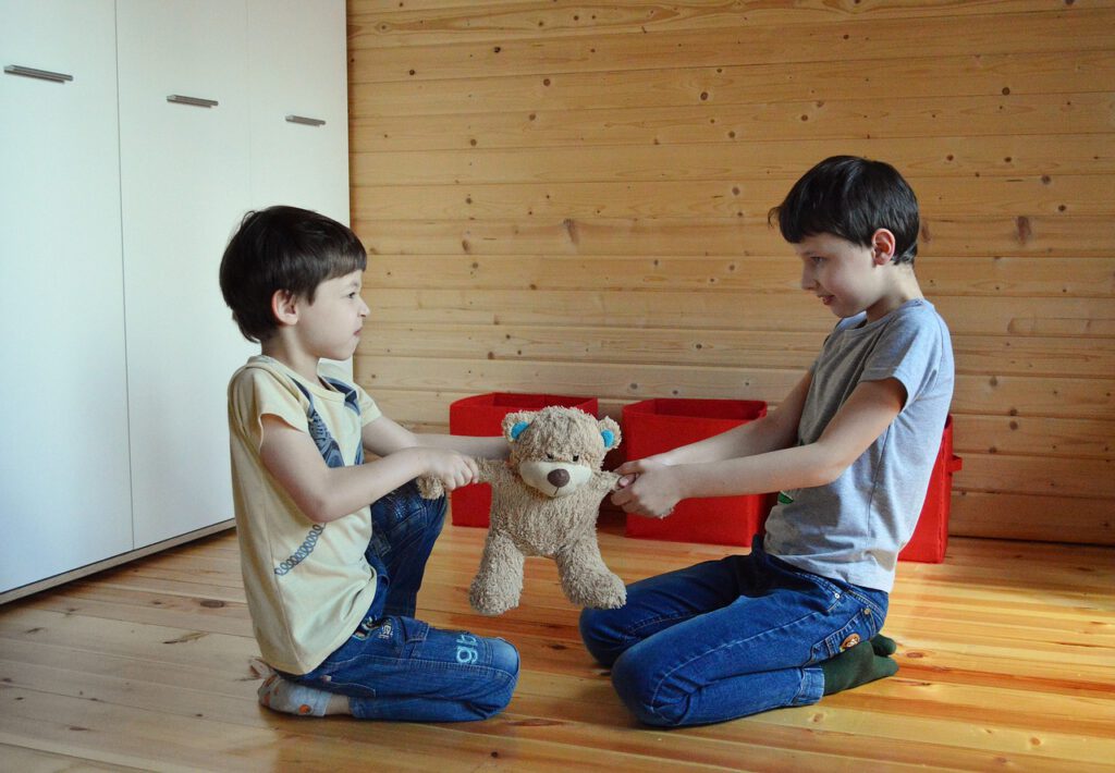 Zwei Kinder sitzen sich auf den Knien gegenüber und zerren an einem Stoff-Teddybären.