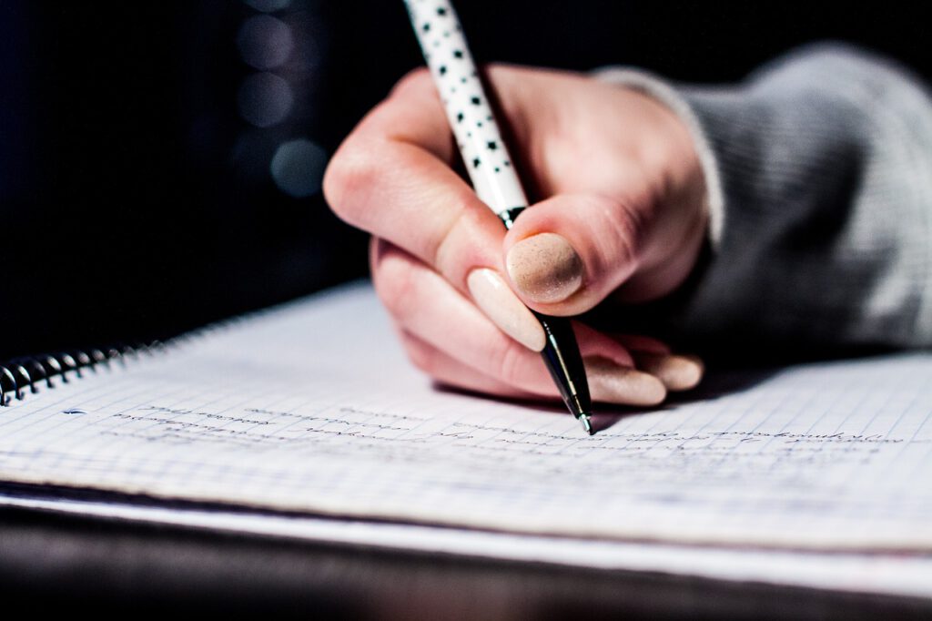 Auf dem Bild sieht man eine Hand, die einen Stift hält und damit auf einem karierten Spiralblock schreibt.