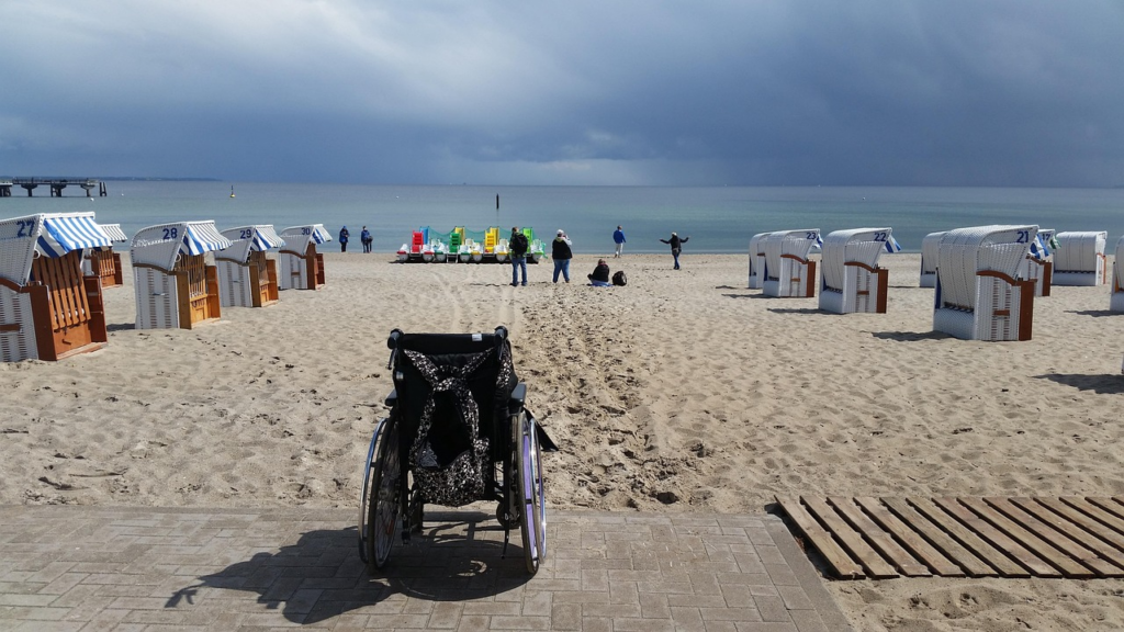 Das Bild zeigt einen Sandstrand mit weißen Strandkörben. Im Vordergrund sieht man einen Holzsteg, auf dem ein Rollstuhl steht, ausgerichtet gen Strand. Im Hintergrund sieht man das blaue Meer leuchten.