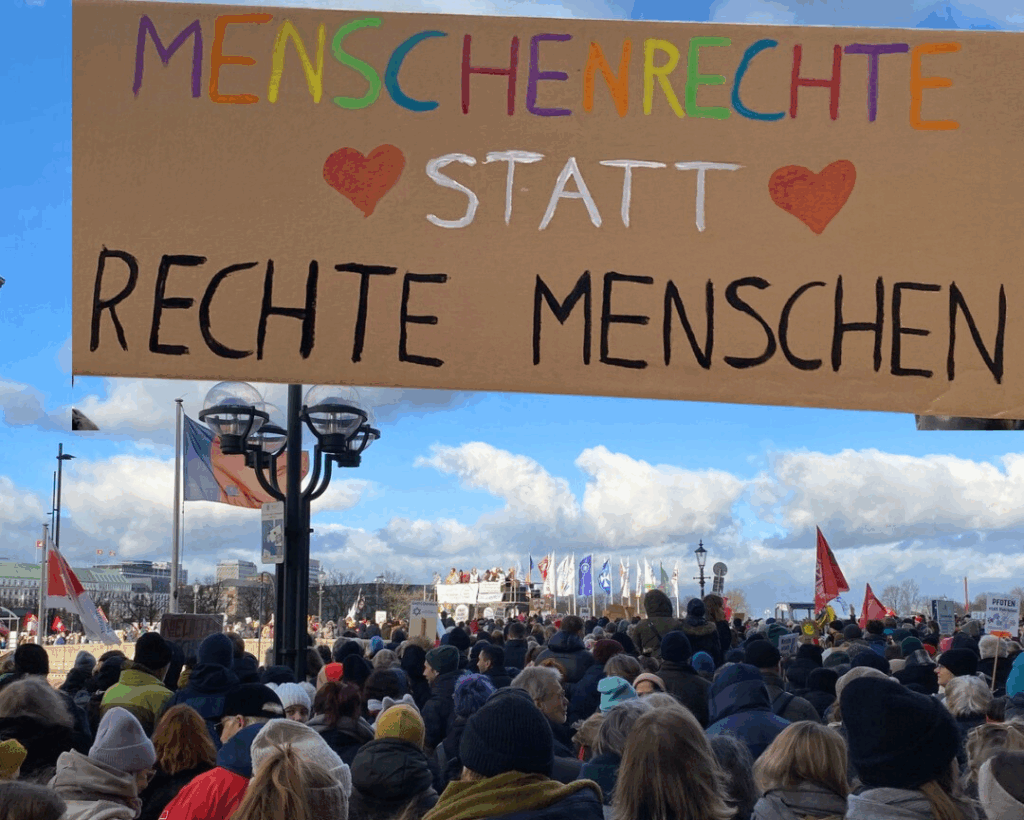 Foto einer großen, protestierenden Menschenmenge am Jungfernstieg. Darüber sieht man ein selbst gemaltes Plakat mit der Aufschrift „Menschenrechte statt rechte Menschen".