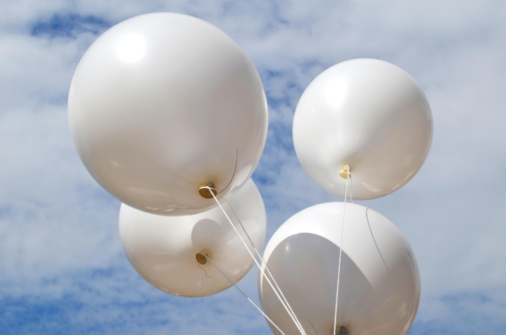 Das Bild zeigt vier weiße Luftballons vor einem blauen Himmel.